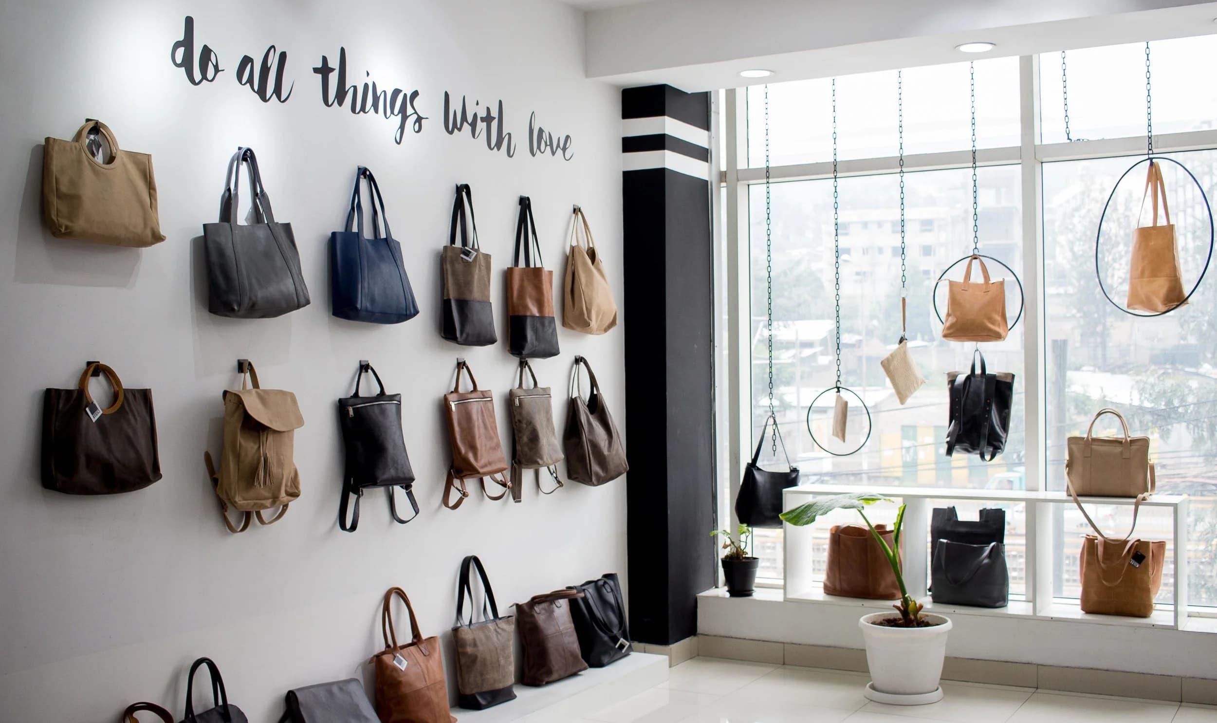 Totes and backpacks hung on a white wall beside a window, under hand-lettered script reading “do all things with love”.