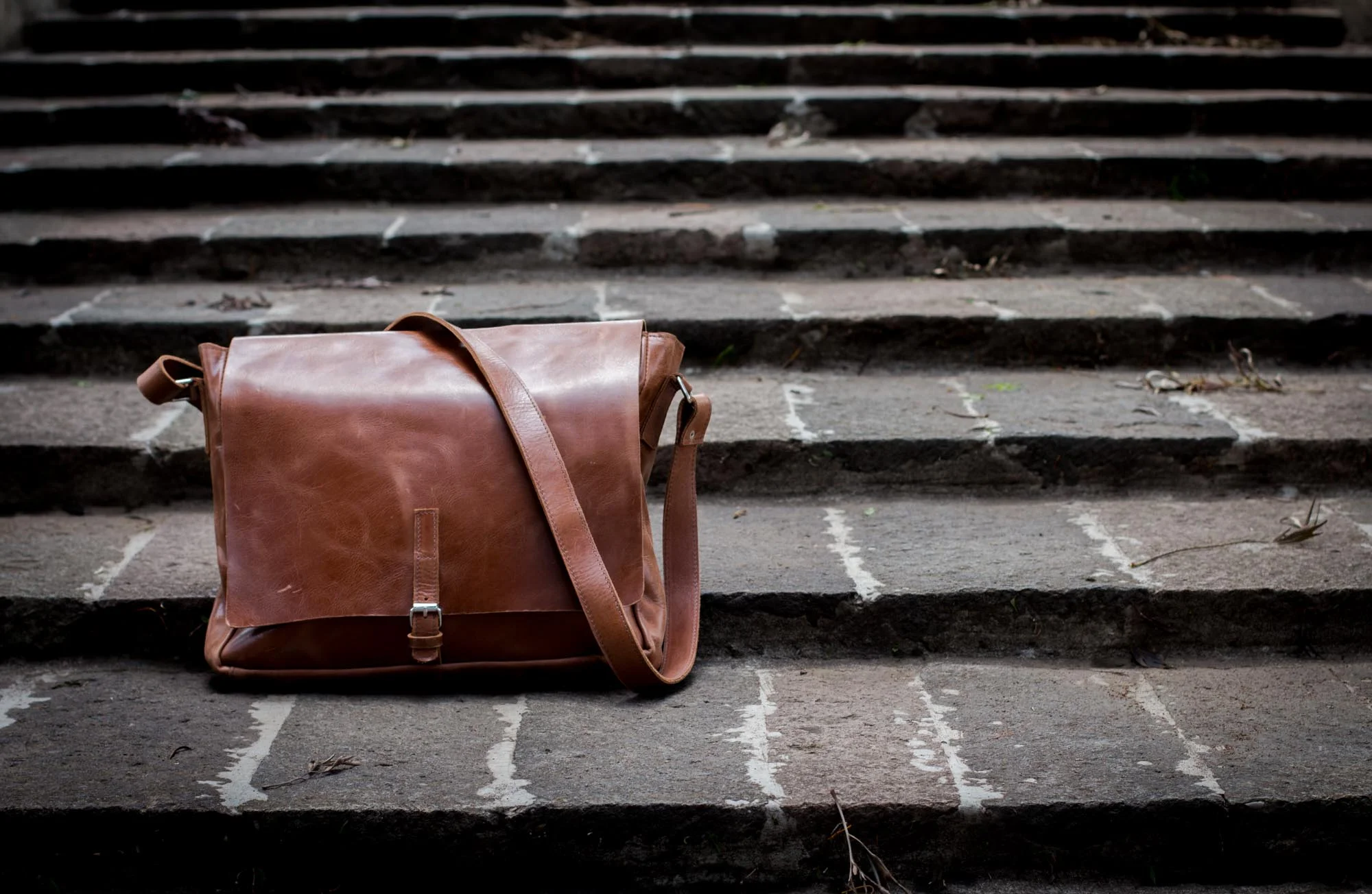 A tan full-grain leather messenger bag resting on worn stone steps.